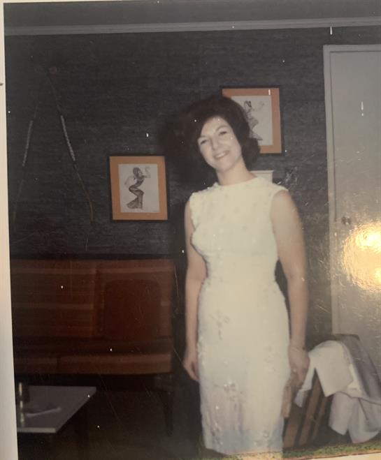 A woman stands smiling in a beautiful white dress in a vintage living room with artwork.