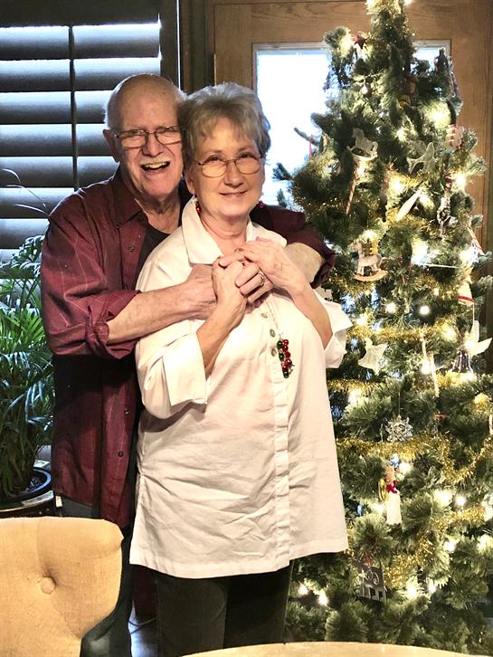 An elderly couple smiles happily together near a Christmas tree adorned with lights.