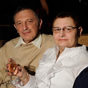 An elderly couple shares a moment at a gathering, both smiling while enjoying cocktails.