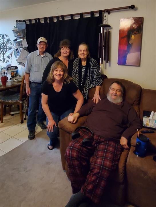 Family members gather around a seated man in a comfortable living room, sharing smiles and laughter.