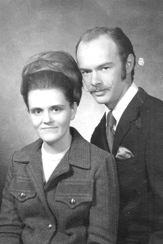 Couple dressed in 1970s formal wear poses together, showcasing elegant hairstyles and outfits.