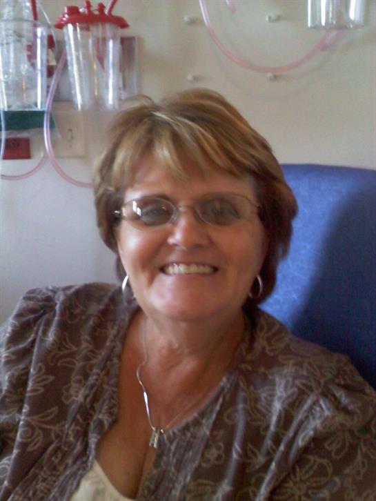A cheerful woman is seated in a hospital room, surrounded by medical apparatus.
