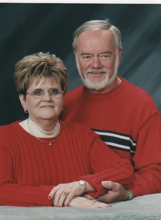 An elderly couple stands close together, dressed in red sweaters, smiling warmly at the camera.