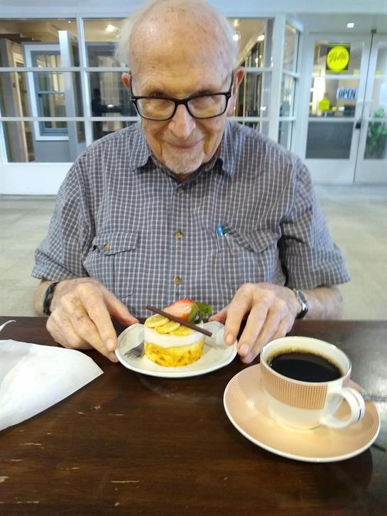 A smiling elderly man sits at a table with a slice of cake and a cup of coffee, enjoying his treat.