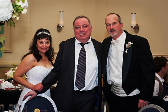 A bride and groom pose with a guest at their wedding reception, smiling and celebrating.