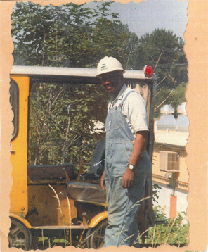A man in blue overalls stands next to a yellow vehicle with buildings and trees in the background.