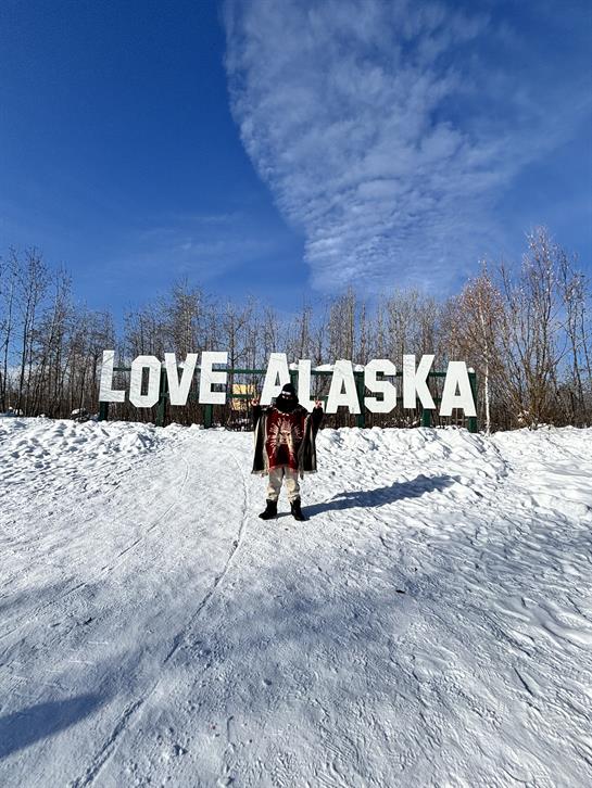 Tourists pose joyfully by the love alaska sign under a bright blue sky on a snowy day.