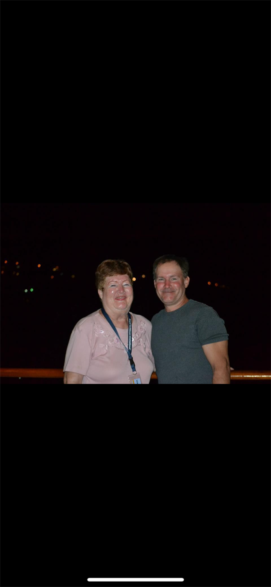 A couple smiles together on a cruise ship deck at night with twinkling lights.