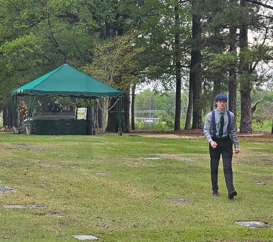 A person in formal clothes walks calmly in a peaceful outdoor setting with a green tent.