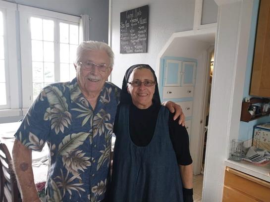 An elderly couple stands close together in a bright kitchen, sharing smiles and warmth.