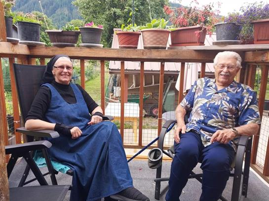 Two older women enjoy each other's company on a sunny porch filled with vibrant plants.