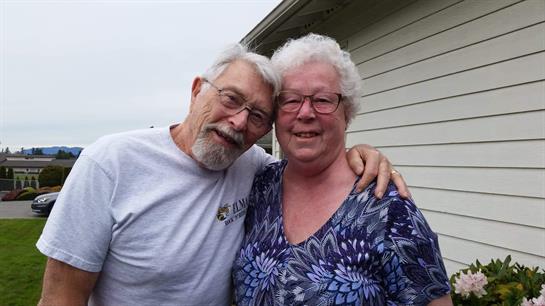 An elderly couple poses happily outdoors, embracing each other in front of their house.