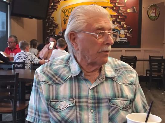 A senior man in a plaid shirt relaxes at a table while others chat and dine around him.