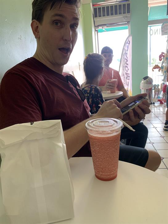 Individual sits at a table, engaged with their phone while savoring a cool drink.