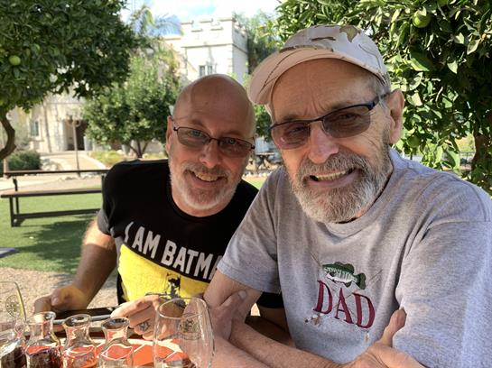 Two friends relax outdoors, sharing drinks and smiles in a lush garden setting.