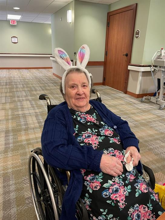 A joyful elderly woman in a wheelchair wears bunny ears at a nursing home celebration.