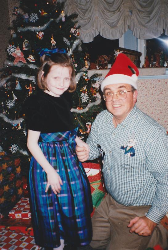 A girl in a plaid dress poses next to a man wearing a Santa hat by a decorated Christmas tree.