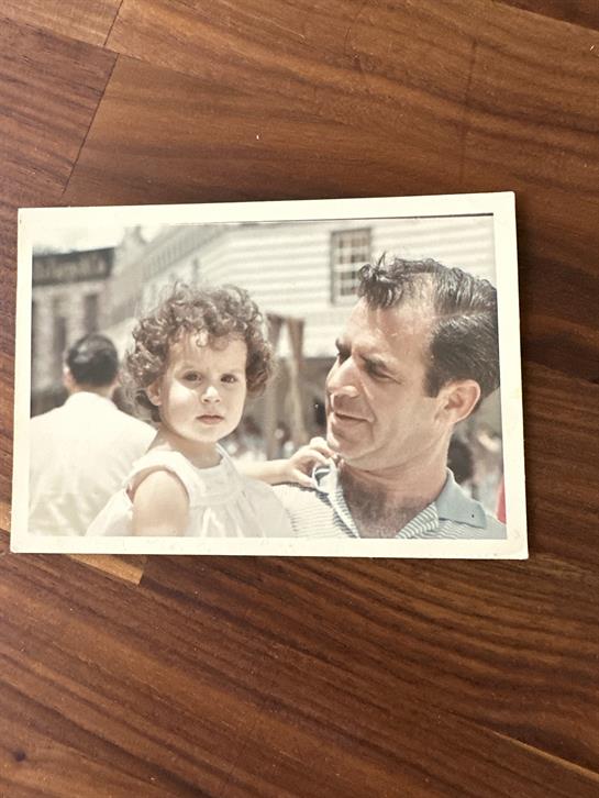 A man holds his daughter, both smiling, while enjoying a warm sunny day outside.
