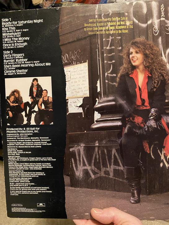 A woman with curly hair sits on steps, holding a music album cover, surrounded by urban art.