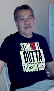 A man poses indoors wearing a shirt that references a high school in Tucson while smiling softly.