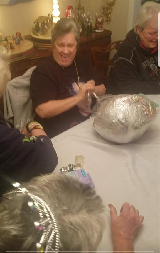 Friends share laughter while unwrapping a surprise gift in a warmly decorated dining area.