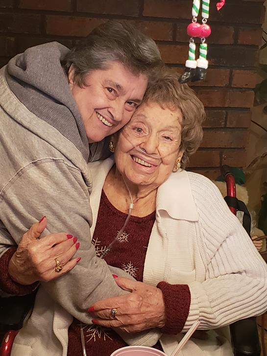 Two smiling women embrace lovingly in a warm living room adorned for the winter festivities.