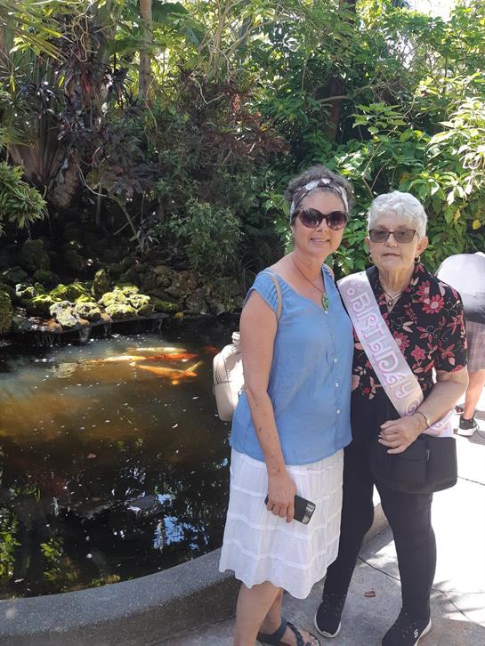 Two women smile together near a serene pond surrounded by vibrant greenery on a sunny day.