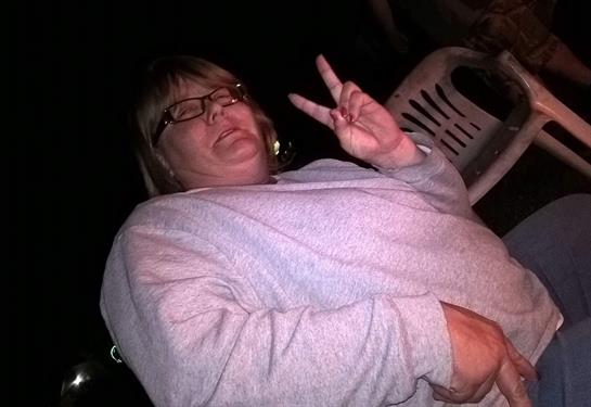 A woman in a gray sweater playfully makes a peace sign while seated outdoors at night.