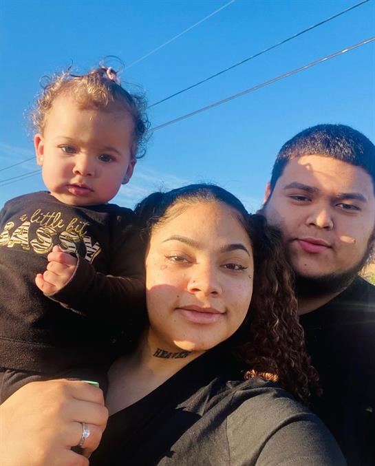 A woman holds a toddler while posing with a man outdoors under a clear blue sky.