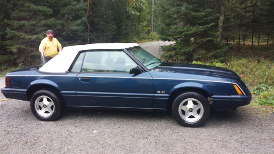 A blue convertible car with a white top is parked on a gravel road surrounded by lush greenery.