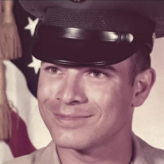 A cheerful soldier in uniform stands confidently with an American flag behind him.