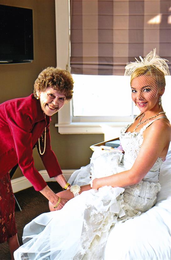 Bride smiles while grandmother adjusts her dress in a warmly lit room before the ceremony.