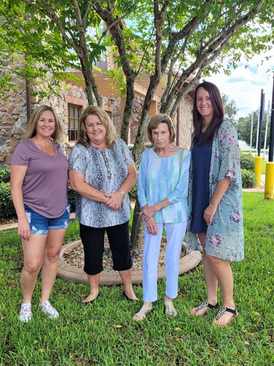 Four women stand together in a park with greenery, enjoying a warm day and smiling joyfully.