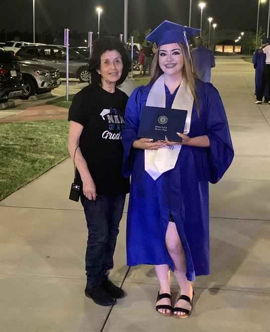 A graduate in cap and gown poses with a family member outdoors, celebrating their achievement.