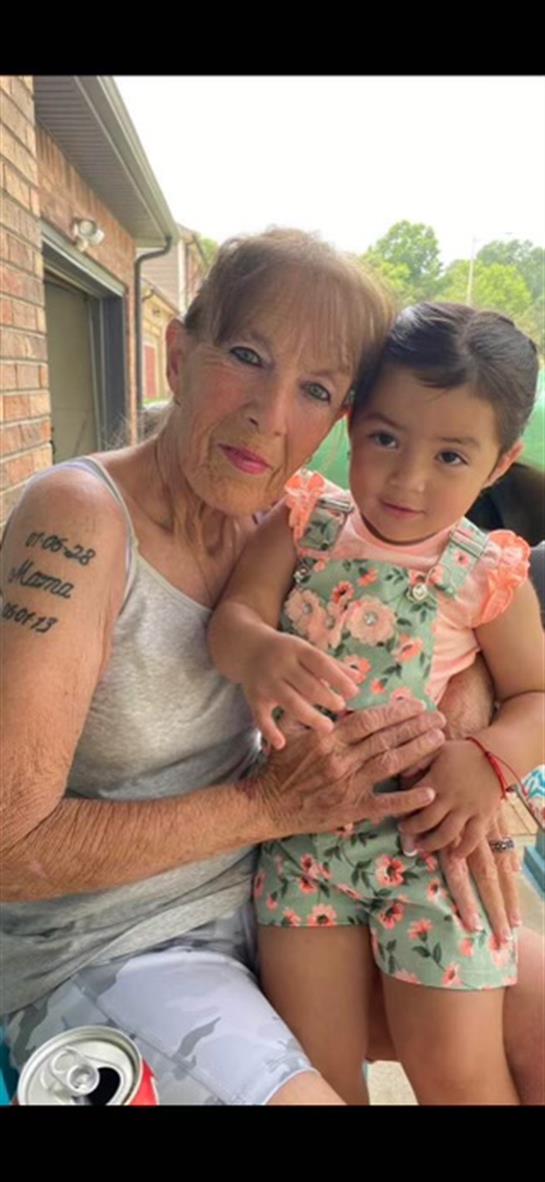 An elderly woman smiles while holding her young granddaughter on a porch in summer.