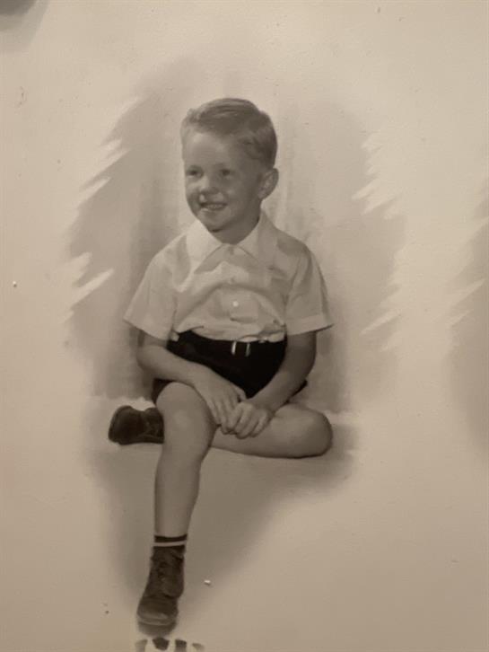 A young boy smiles while sitting with his legs crossed in a modest environment.