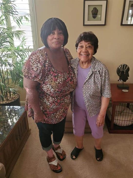 Two women smile together in a warm living room filled with natural light and decorations.