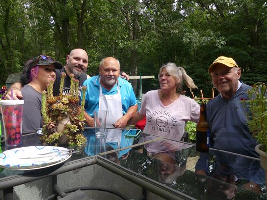 Five friends share drinks and laughter at a lively barbecue in a green backyard.