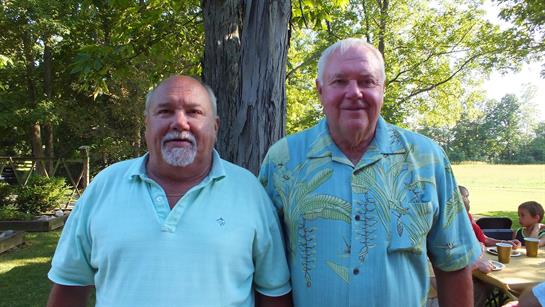 Two men stand side by side smiling in a green park setting under a large tree.