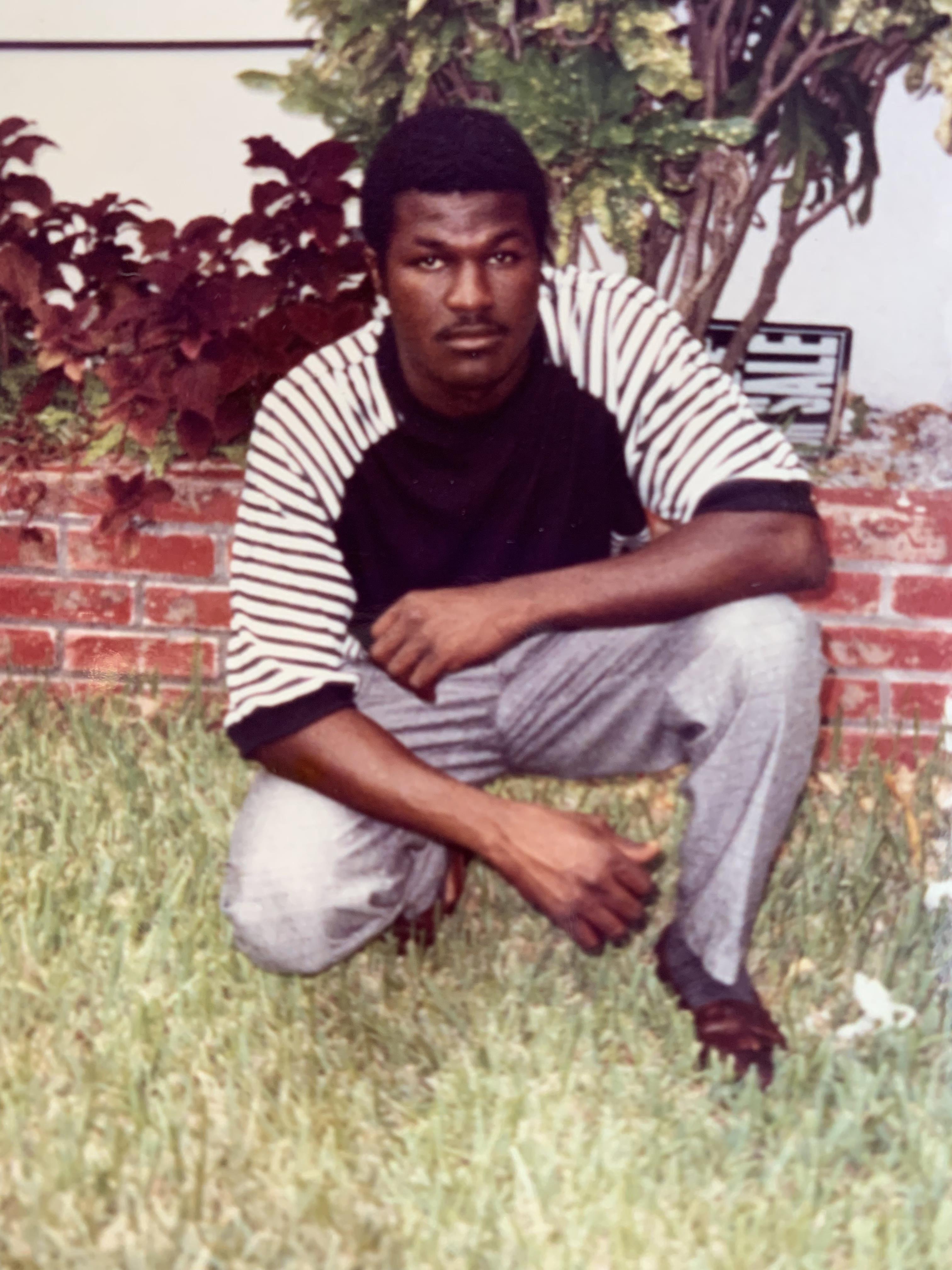 A man with a serious expression crouches on grass, surrounded by greenery and a brick wall.