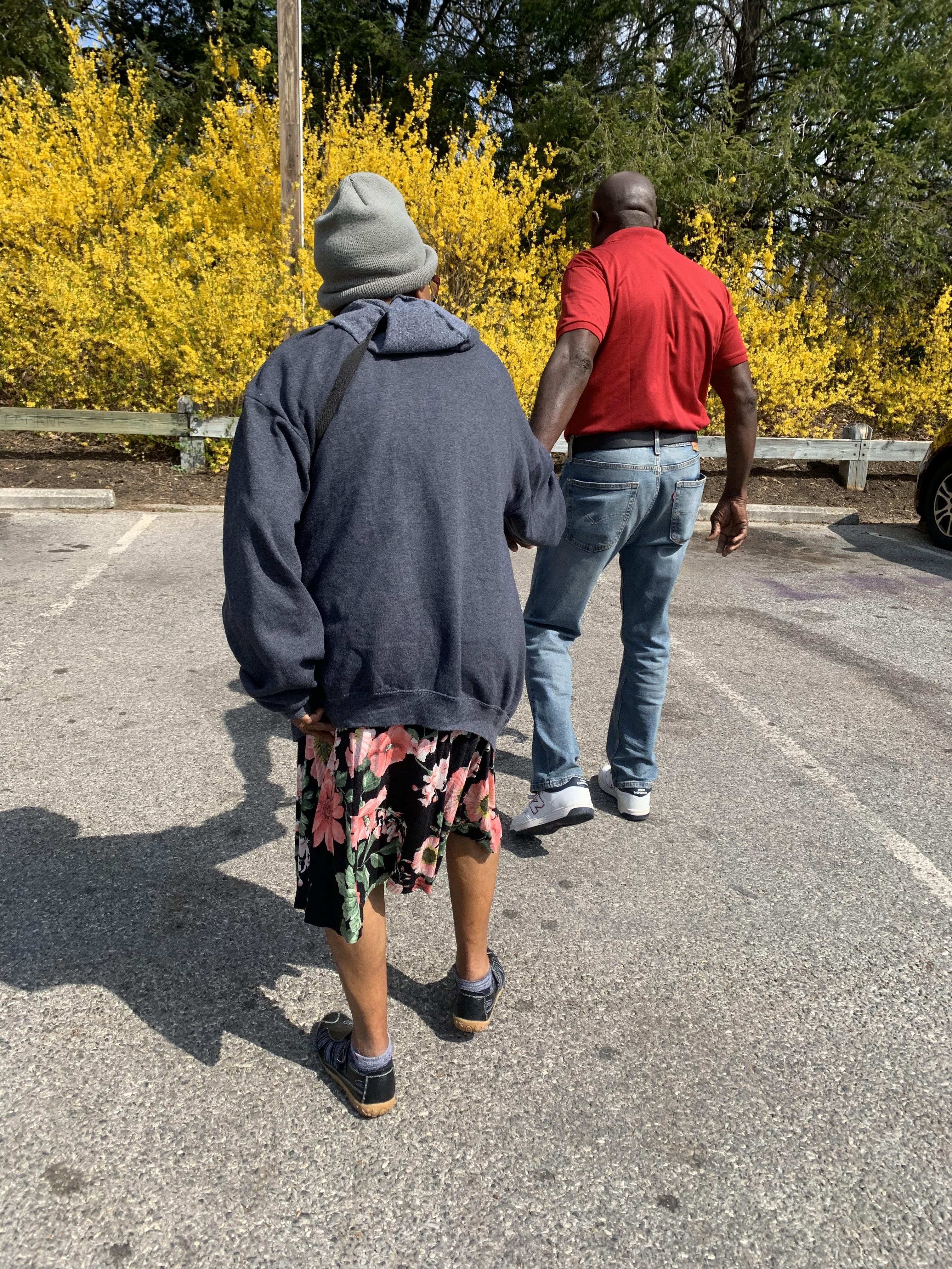 Two people stroll in a sunny parking lot with bright yellow flowers nearby.