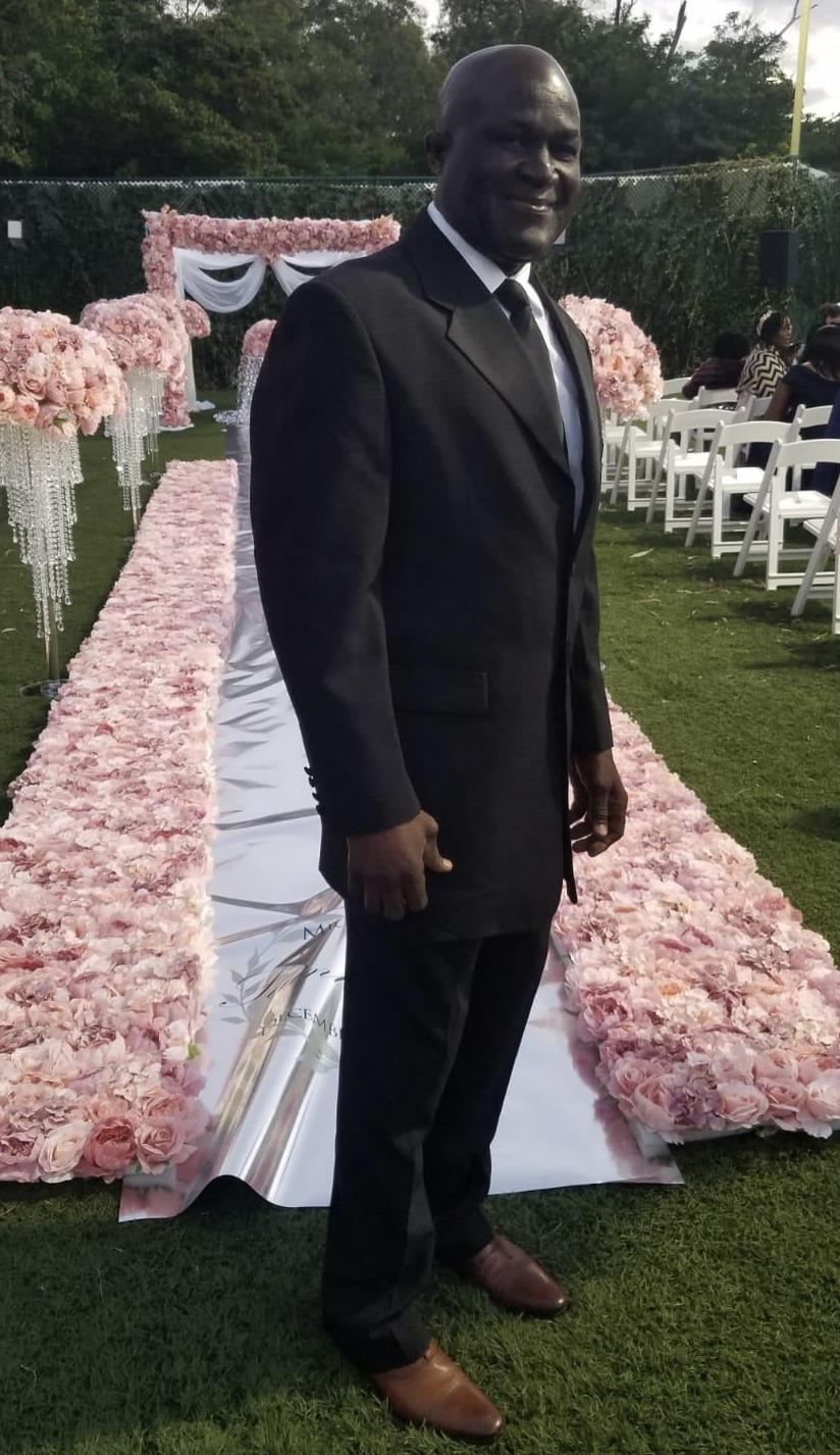 A well-dressed man poses at an outdoor wedding with pink floral decorations lining the aisle.