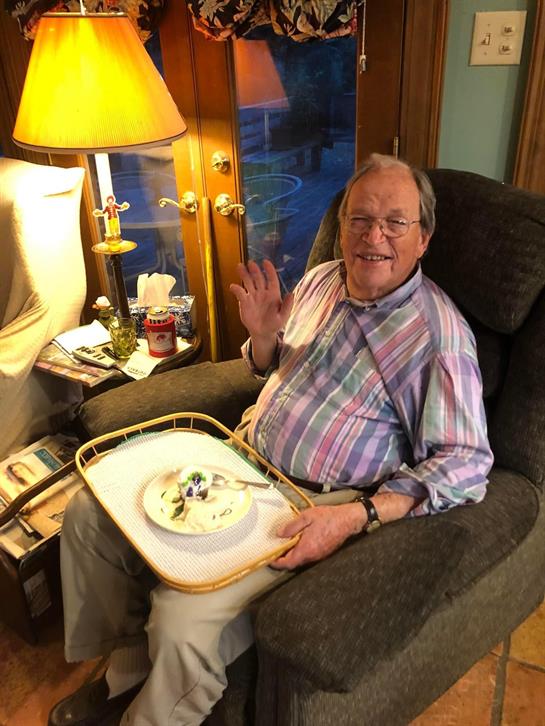 A cheerful elderly man sits in an armchair, waving while holding a tray with food.