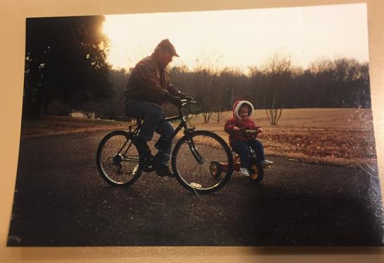 A man rides a bicycle alongside a child on a tricycle in a peaceful park setting.