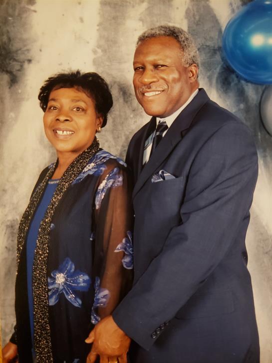 Couple dressed in formal wear smiles together against a decorative background with balloons.