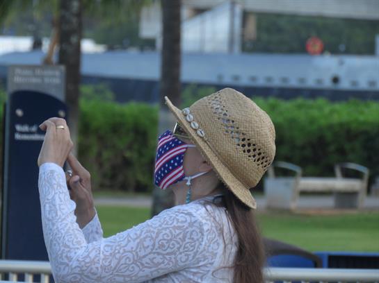 A woman in a straw hat and mask is capturing a selfie in a green park area.