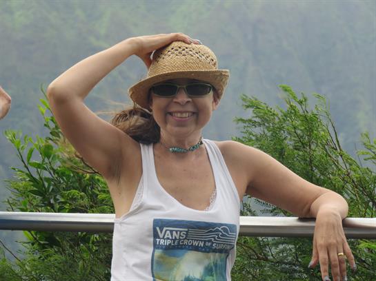 A woman smiles while resting against a railing surrounded by vibrant green plants.