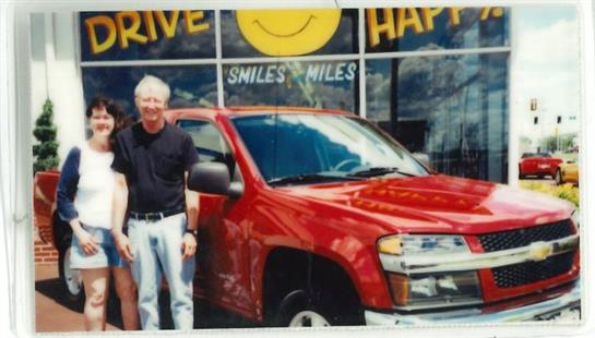 A couple stands proudly beside their new red truck in front of a bright dealership.