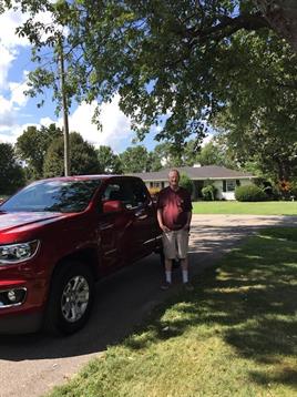 An elderly man poses near a bright red truck on a sunny afternoon in a quiet area.
