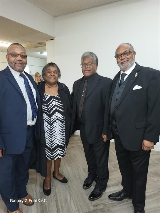 Four individuals dressed in formal attire pose together and smile at a celebration indoors.